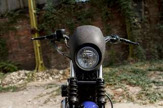 Harley-Davidson 883 Iron — Frontale: cupolino nero, faro, manubrio, ghiaia e strutture in legno alle spalle.