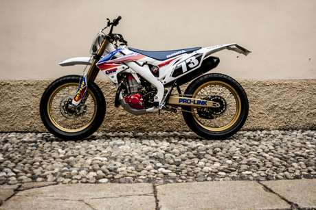 Honda CRF 450 E — Fiancata sinistra completa all'aperto, muro chiaro con zoccolo di ciottoli.