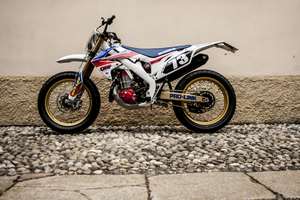 Honda CRF 450 E — Fiancata sinistra completa all'aperto, muro chiaro con zoccolo di ciottoli.